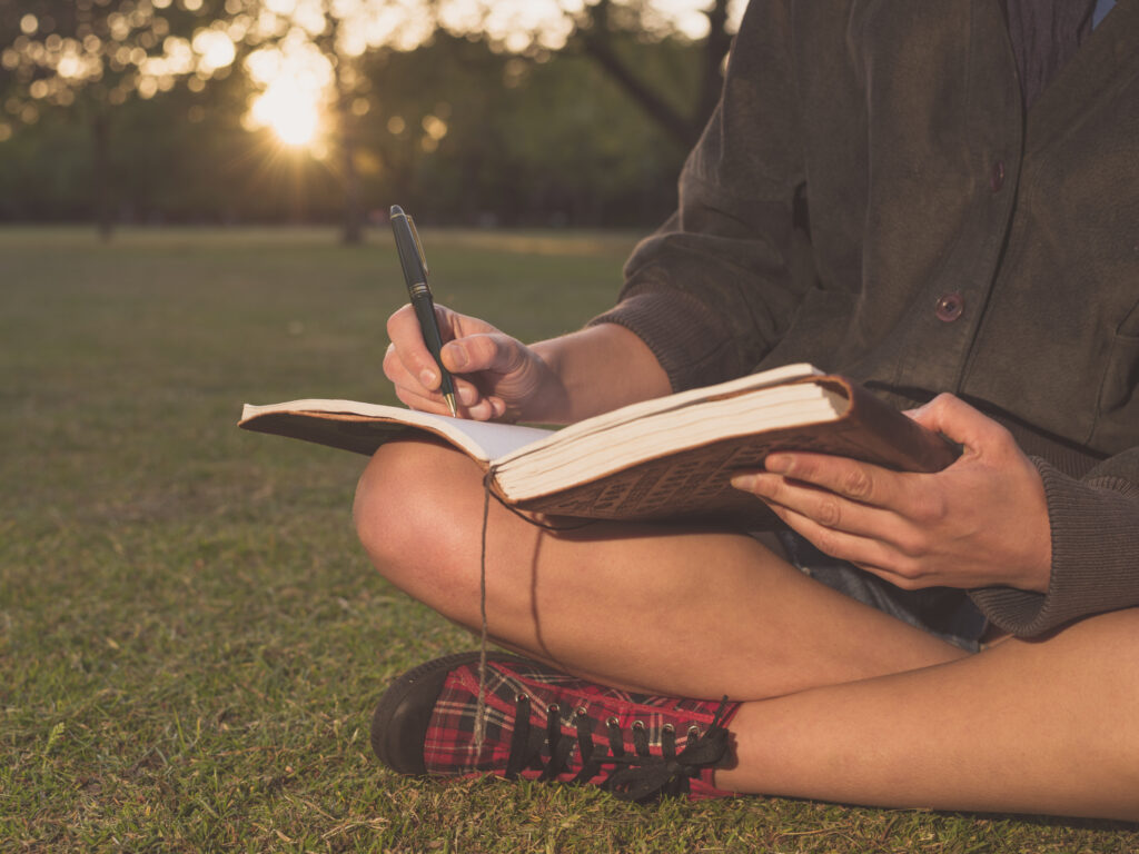 A person, viewed from the shoulders down, is wearing red plaid sneakers, a dark shirt and shorts. They are sitting cross legged on the grass at sunset and writing with a pen in a journal.